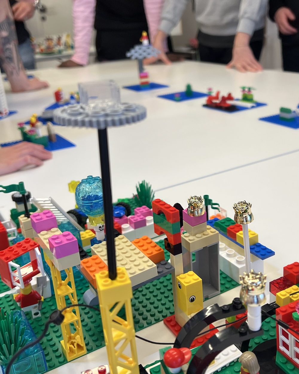 Modèle collectif en briques LEGO® SERIOUS PLAY® sur plaques vertes, grue jaune, couronnes dorées et engrenage en haut d’un mât, et derrière, des modèles individuels posés chacun sur une plaque bleue, entourés des mains des participants